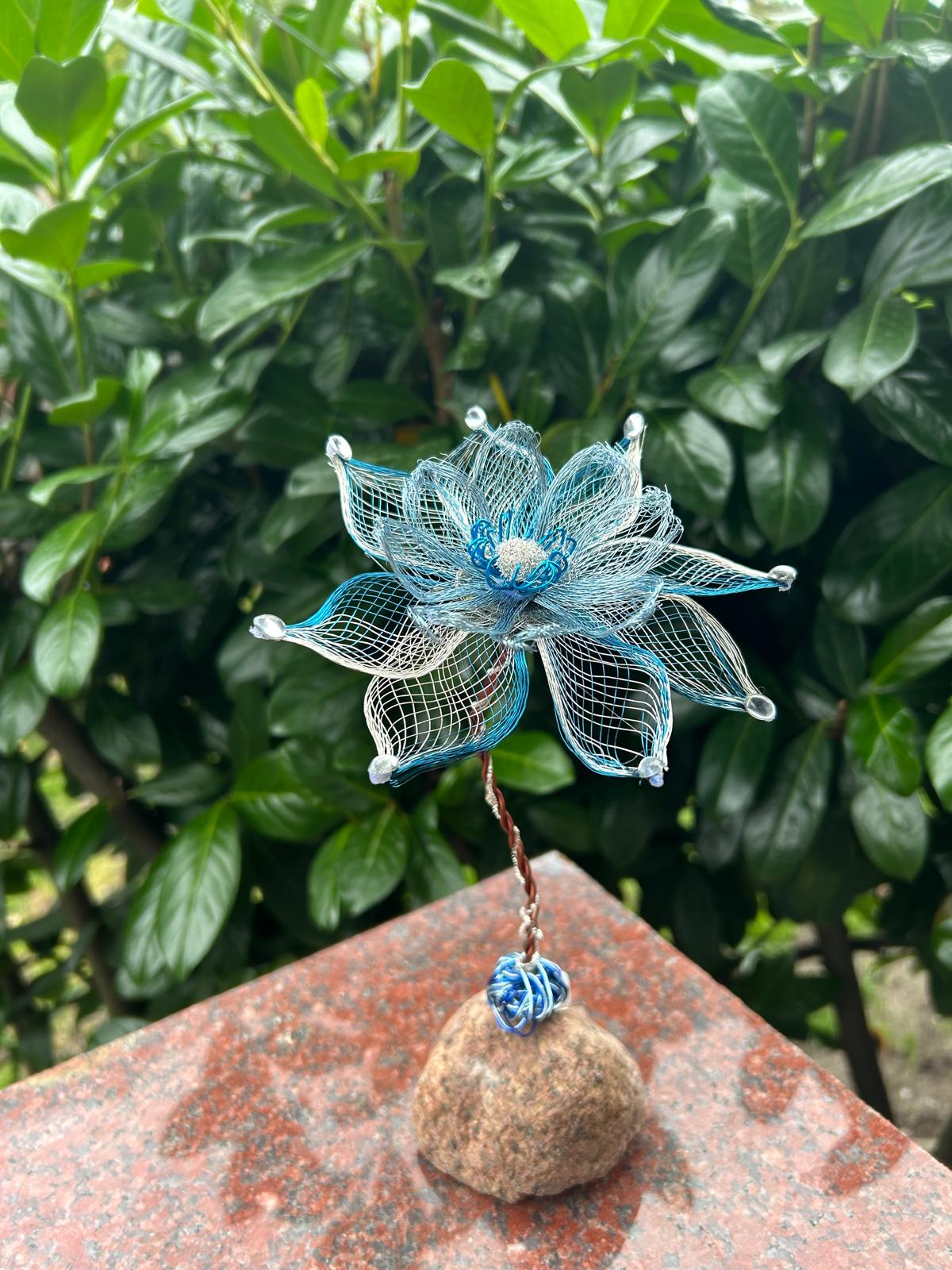 Blau-silberne Drahtblüte mit kleinen Perlen an den Blattspitzen auf einem Naturstein vor grünem Laub