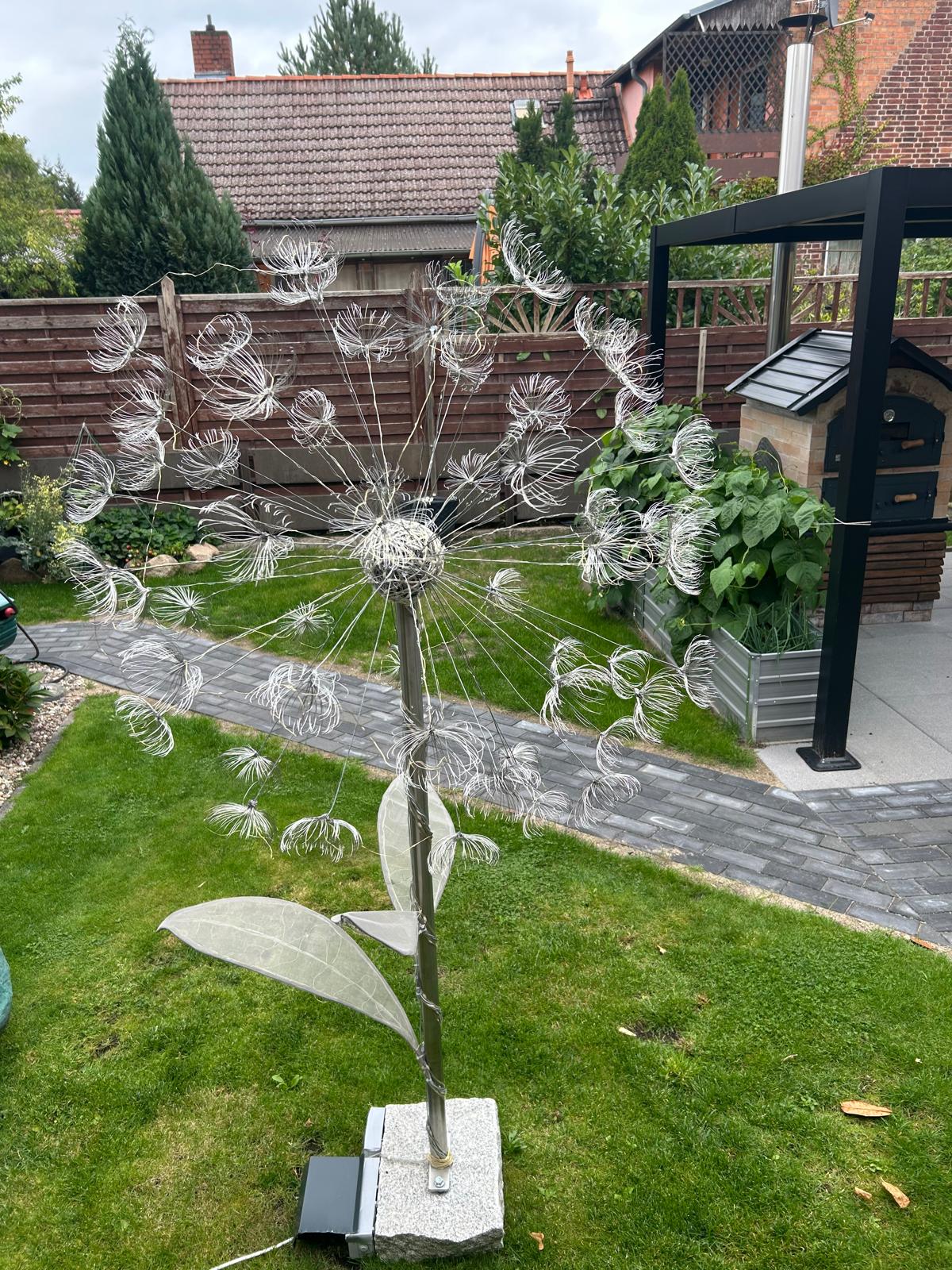Große silberne Drahtpusteblume mit vielen gebogenen Schirmchen und Gewebeblättern auf einem Granitsockel im Garten