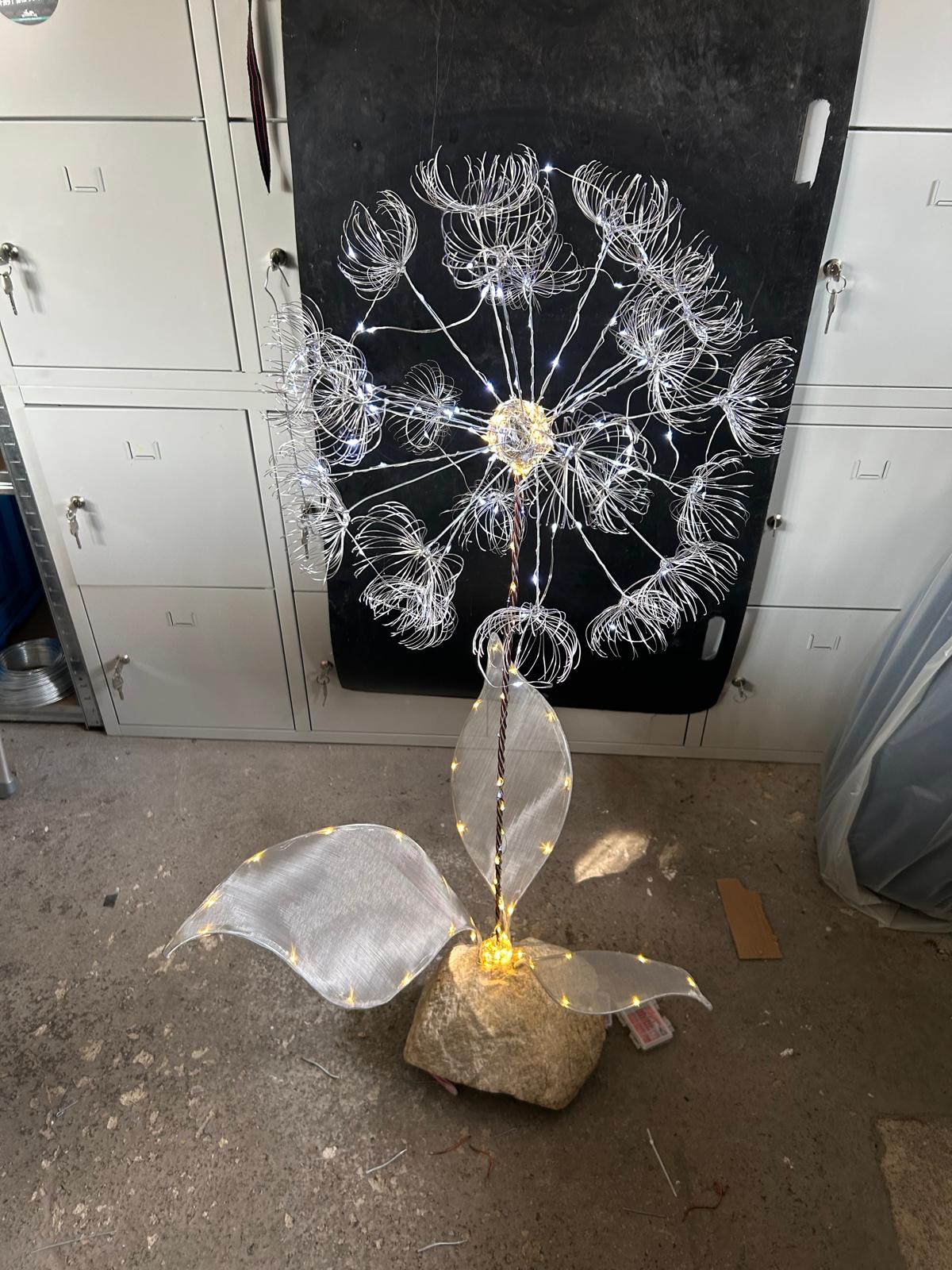Silberne Drahtpusteblume mit beleuchteten Schirmchen und großen Gewebeblättern auf einem Naturstein