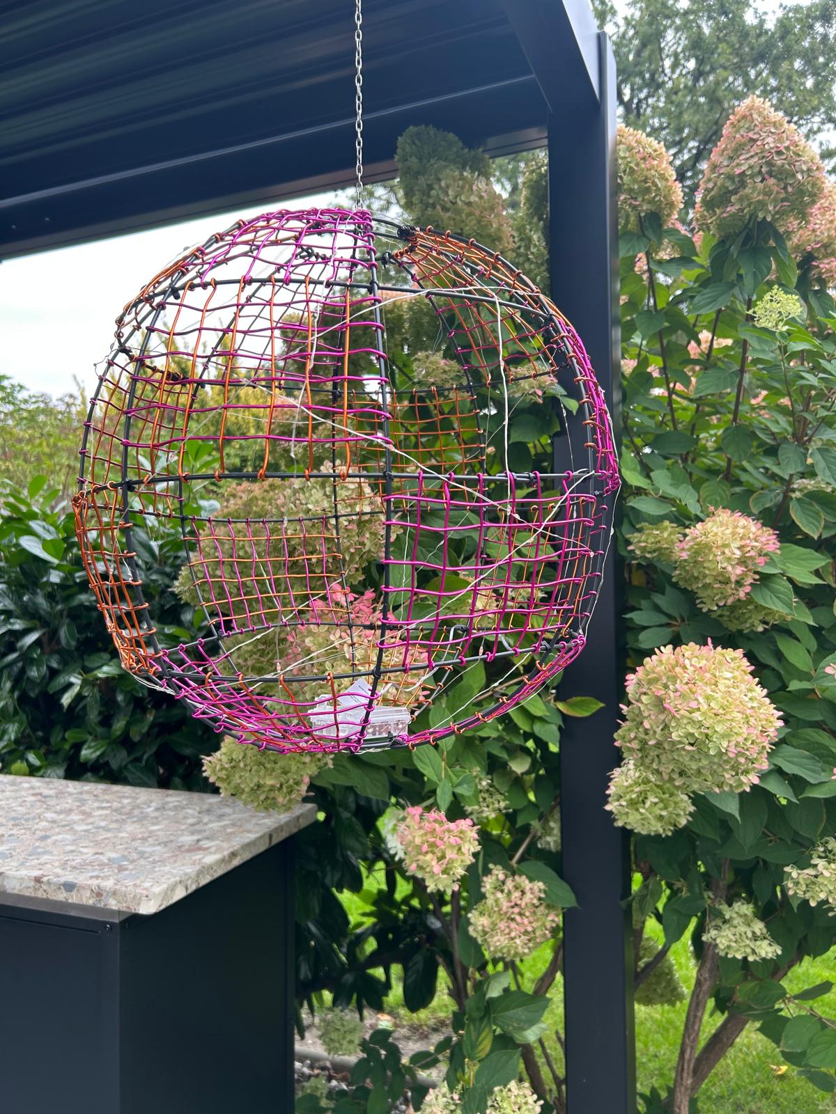 Magenta-orange Drahtweltkugel an einer Kette vor blühenden Hortensien